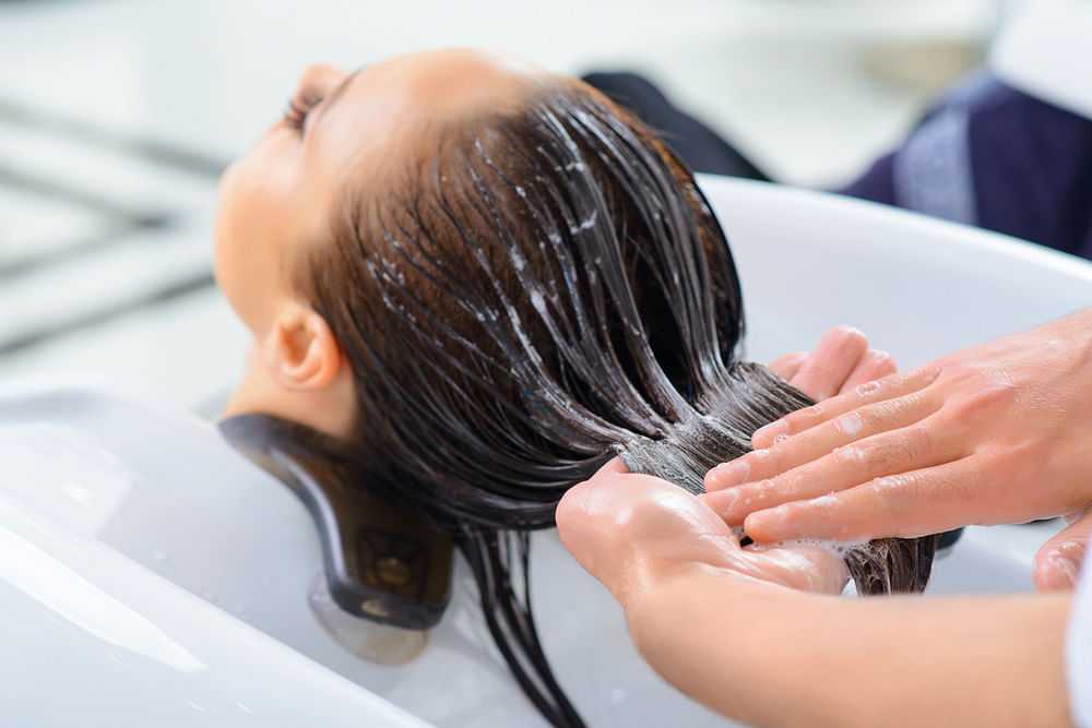 Hair being washed at a salon with hands massaging shampoo into long, wet hair.