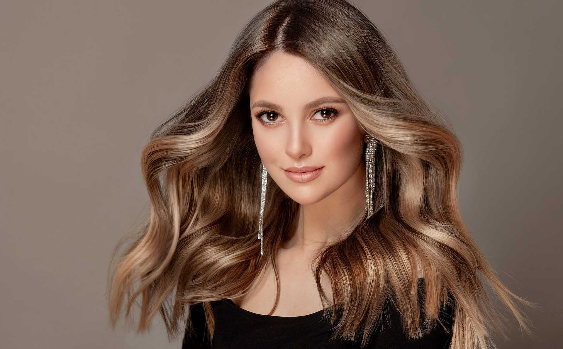 Woman with long, flowing hair and elegant earrings in a black top against a gray background.