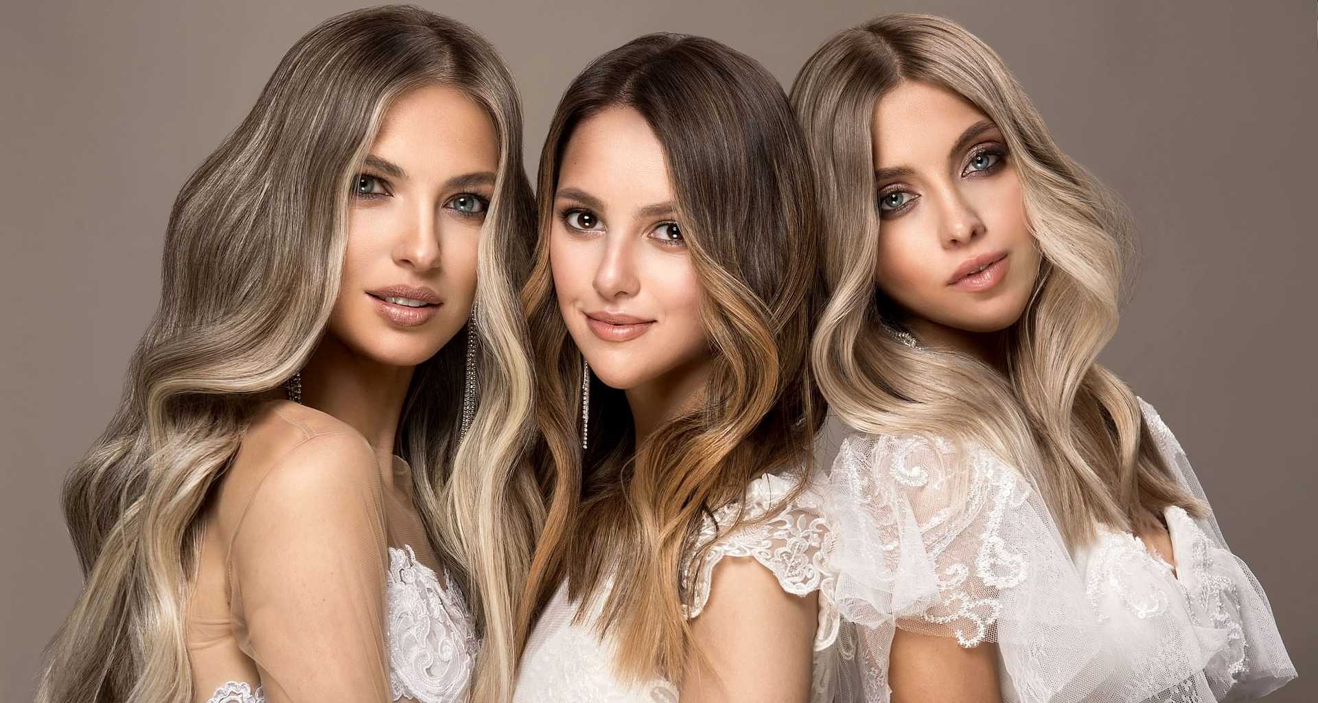 Three women with wavy hair in white dresses pose against a neutral backdrop.