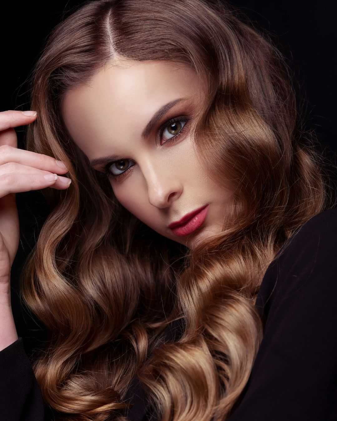 Elegant woman with wavy brunette hair and soft makeup against a dark background.