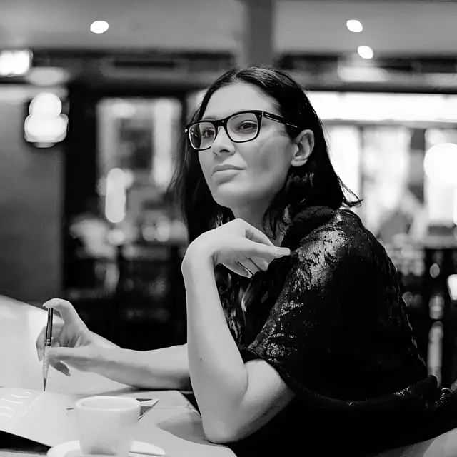 Woman sitting at a café with a notebook and pen, wearing glasses and looking thoughtful.
