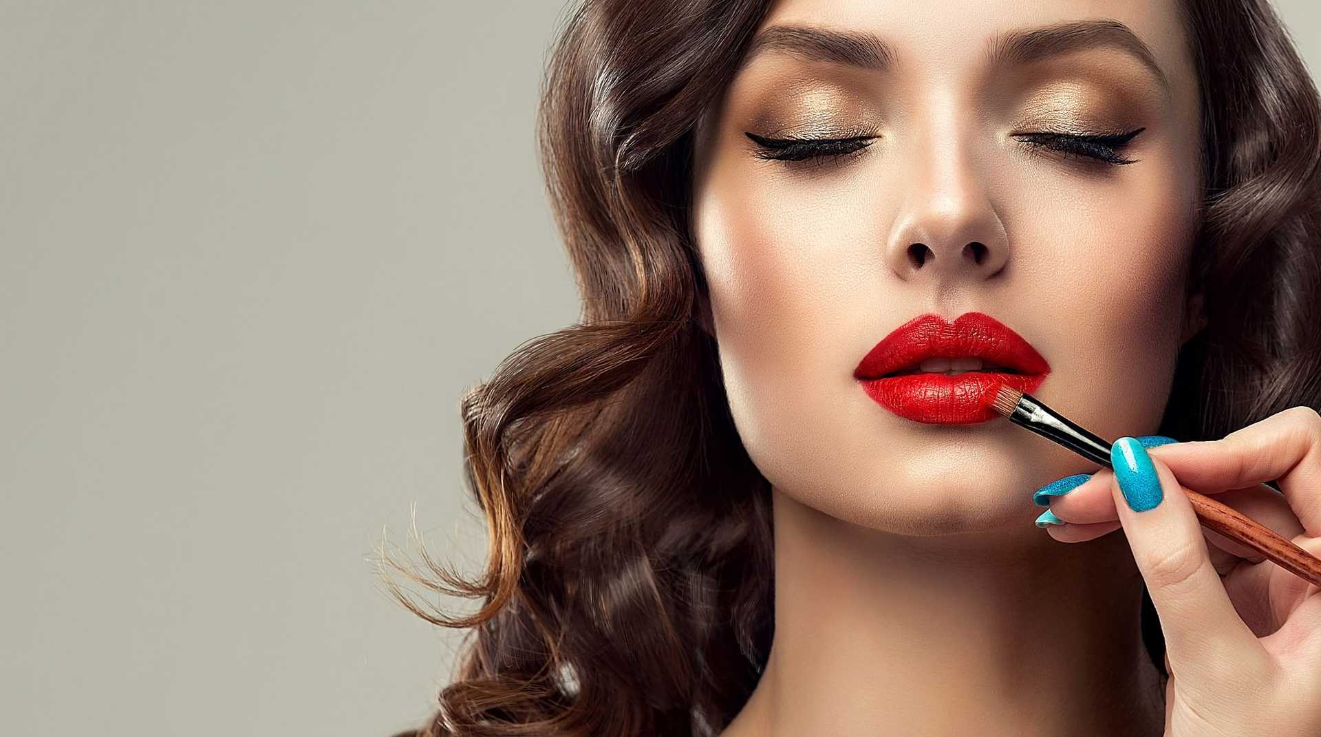 Woman's lips being painted with vibrant red lipstick by a makeup artist.